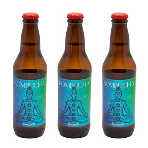 La imagen muestra tres botellas ámbar de Kombucha Kombox alineadas horizontalmente. Cada botella tiene una tapa roja y una etiqueta azul con la imagen de una persona en posición de loto y los chakras alineados. El fondo es neutro y claro.