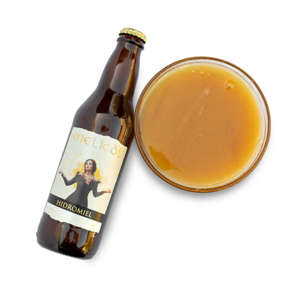 La imagen presenta una botella de hidromiel Melicol inclinada hacia un lado, con su etiqueta amarilla y beige mostrando una mujer con los brazos extendidos. Al lado, un vaso lleno de miel polifloral de color ámbar claro, todo sobre un fondo blanco que resalta la bebida y la botella.