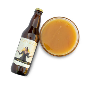 La imagen presenta una botella de hidromiel Melicol inclinada hacia un lado, con su etiqueta amarilla y beige mostrando una mujer con los brazos extendidos. Al lado, un vaso lleno de miel polifloral de color ámbar claro, todo sobre un fondo blanco que resalta la bebida y la botella.