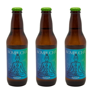 La imagen muestra tres botellas ámbar de Kombucha Kombox alineadas horizontalmente. Cada botella tiene una tapa verde y una etiqueta azul con la imagen de una persona en posición de loto y los chakras alineados. El fondo es neutro y claro.